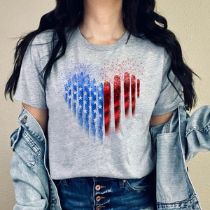 Pu&ograve; includere: Una maglietta grigia con un design a forma di cuore a tema bandiera americana. Il design incorpora stelle blu e strisce rosse e bianche, creando una rappresentazione visiva della bandiera.