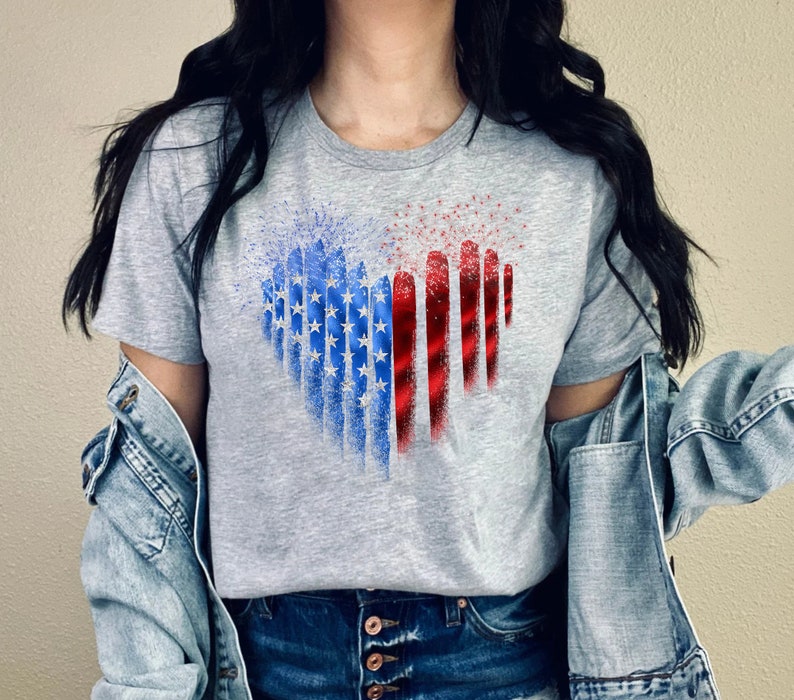 Pu&ograve; includere: Una maglietta grigia con un design a forma di cuore a tema bandiera americana. Il design incorpora stelle blu e strisce rosse e bianche, creando una rappresentazione visiva della bandiera.