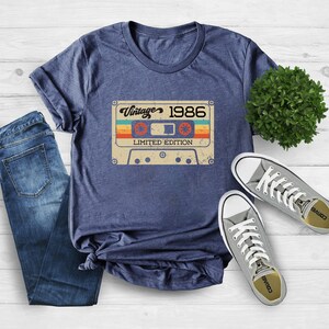 May include: A navy blue t-shirt featuring a vintage cassette tape graphic. The tape has the text "Vintage 1986 Limited Edition" printed on it.
