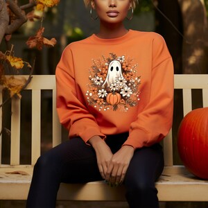 May include: Orange sweatshirt featuring a white ghost illustration surrounded by flowers and pumpkins. The design is centered on the front of the long-sleeved top. The sweatshirt is a solid orange color, perfect for Halloween or fall.