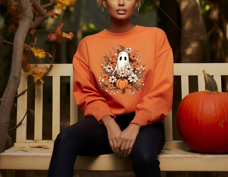May include: Orange sweatshirt featuring a white ghost illustration surrounded by flowers and pumpkins. The design is centered on the front of the long-sleeved top. The sweatshirt is a solid orange color, perfect for Halloween or fall.