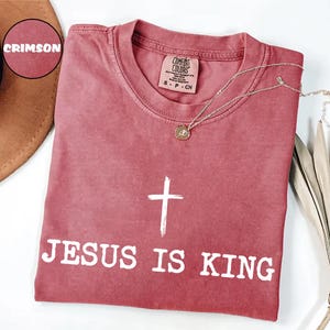 K&ouml;nnte beinhalten: Karmesinrotes T-Shirt mit dem Text "JESUS IS KING" und einer wei&szlig;en Kreuzgrafik. Das Shirt hat einen Rundhalsausschnitt und ein kleines Etikett am Ausschnitt. Eine goldene Halskette mit Anh&auml;nger liegt &uuml;ber dem Shirt.