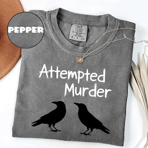 May include: A gray t-shirt with the words "Attempted Murder" and two black crow silhouettes. The shirt has a crew neck and a small tag. A silver necklace with a pendant is draped on the shirt.