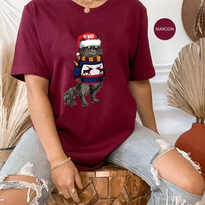 K&ouml;nnte beinhalten: Ein maroonfarbenes T-Shirt mit einem Aufdruck eines Katers, der einen Weihnachtsmann-Hut und einen Pullover mit Rentieren tr&auml;gt. Der Text "My Mode" steht auf dem Hut.