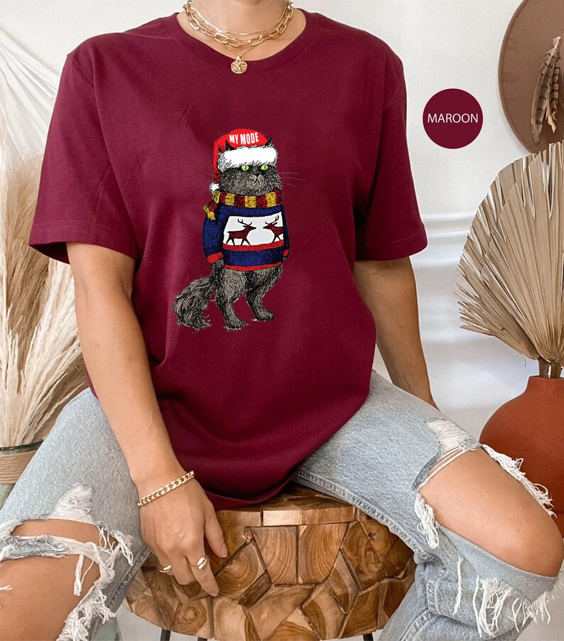 K&ouml;nnte beinhalten: Ein maroonfarbenes T-Shirt mit einem Aufdruck eines Katers, der einen Weihnachtsmann-Hut und einen Pullover mit Rentieren tr&auml;gt. Der Text "My Mode" steht auf dem Hut.