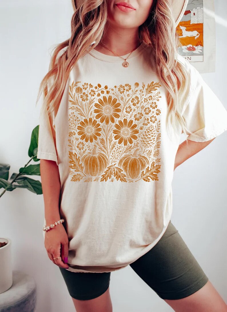 May include: Cream-colored t-shirt featuring a floral and pumpkin design in shades of orange and brown. The graphic includes large sunflowers, pumpkins, and various floral elements. The shirt has short sleeves and a relaxed fit.