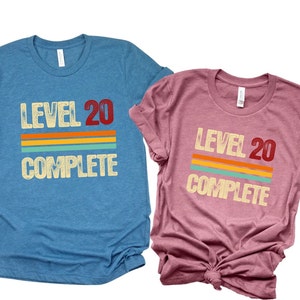 May include: Two t-shirts with a retro design. The blue shirt says "Level 20 Complete" and the pink shirt says "Level 20 Complete".