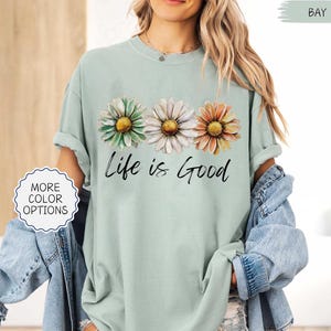 May include: A light green t-shirt with three daisy flowers in green, white, and orange. The words "Life is Good" are written in black script below the flowers. The shirt has rolled-up sleeves.