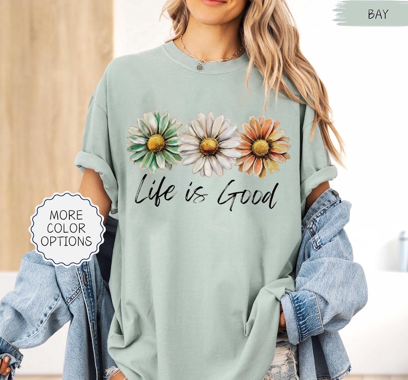 May include: A light green t-shirt with three daisy flowers in green, white, and orange. The words "Life is Good" are written in black script below the flowers. The shirt has rolled-up sleeves.
