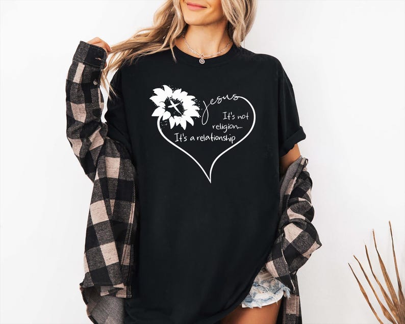 May include: A black t-shirt featuring a white graphic of a sunflower with a cross in the center, shaped into a heart outline. The text reads: Jesus It's not religion... It's a relationship. The shirt is worn with a black and tan plaid button-down shirt.