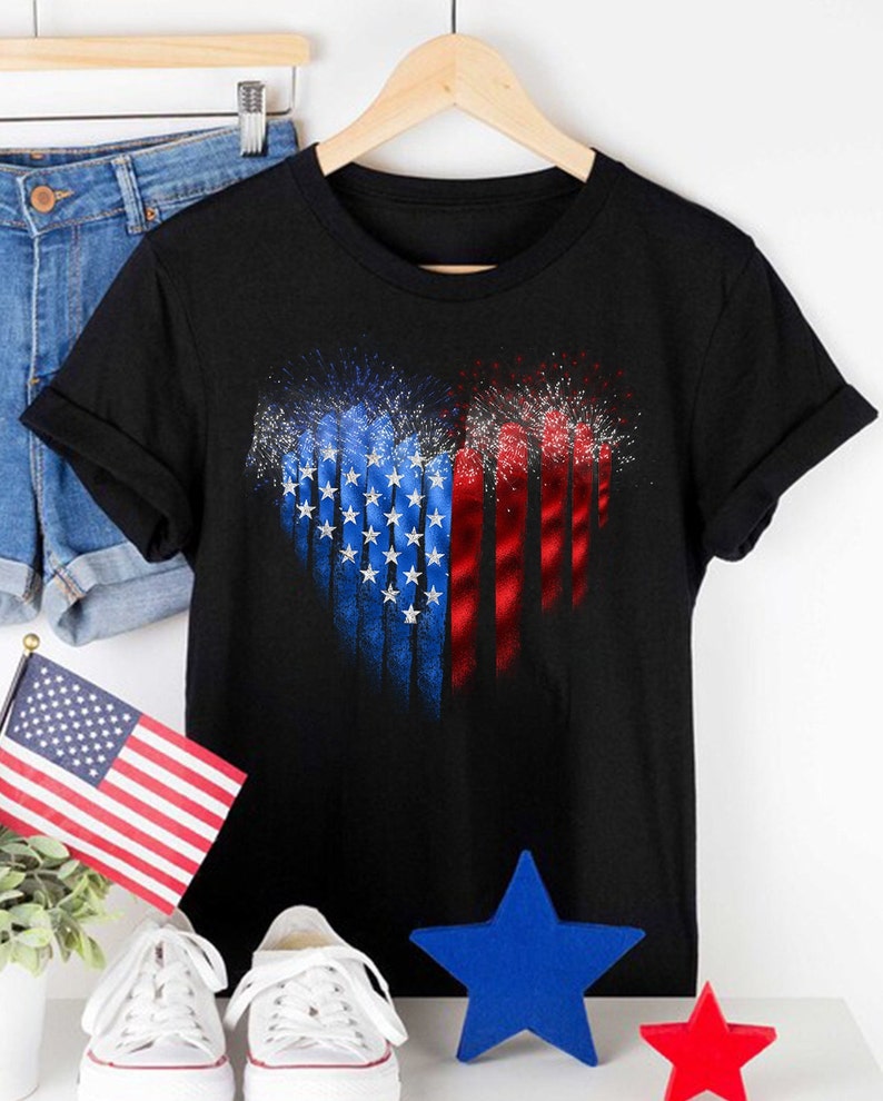 Pu&ograve; includere: T-shirt nero con un design a forma di cuore della bandiera americana e fuochi d'artificio. Il design utilizza blu, rosso e bianco. La maglietta &egrave; esposta con pantaloncini di jeans, una bandiera e decorazioni patriottiche.