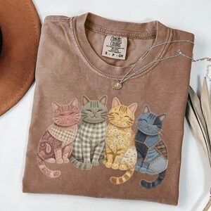 May include: A brown Comfort Colors t-shirt featuring four smiling cat appliqu&eacute;s in various patterns and colors. The cats are arranged in a row, with a paisley, plaid, floral, and patchwork design. A silver necklace with a pendant is draped on the shirt.