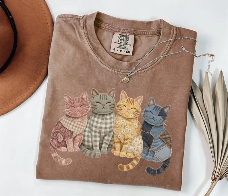 May include: A brown Comfort Colors t-shirt featuring four smiling cat appliqu&eacute;s in various patterns and colors. The cats are arranged in a row, with a paisley, plaid, floral, and patchwork design. A silver necklace with a pendant is draped on the shirt.