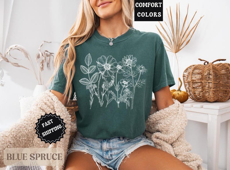 May include: A teal-colored t-shirt with a white floral design. The shirt has short sleeves and a crew neck. The words "COMFORT COLORS" are in a black box in the upper right corner. The words "FAST SHIPPING" are in a black starburst.