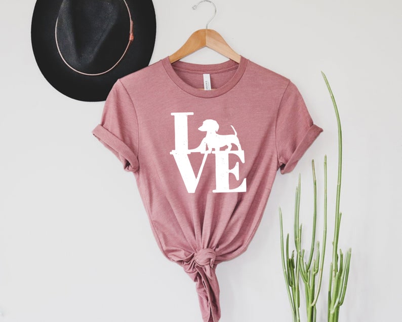 以下が含まれることがあります： マーブ色のTシャツに、白いダックスフントのシルエットが「LOVE」の文字の中に描かれたグラフィックがプリントされています。