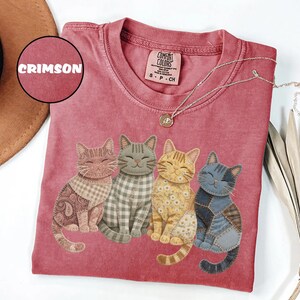 May include: Crimson-colored t-shirt featuring four smiling cat illustrations with patchwork designs. The cats are in various colors and patterns, including paisley, gingham, and floral. The shirt has a crew neck and a Comfort Colors tag.