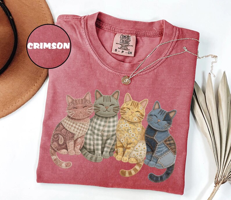 May include: Crimson-colored t-shirt featuring four smiling cat illustrations with patchwork designs. The cats are in various colors and patterns, including paisley, gingham, and floral. The shirt has a crew neck and a Comfort Colors tag.
