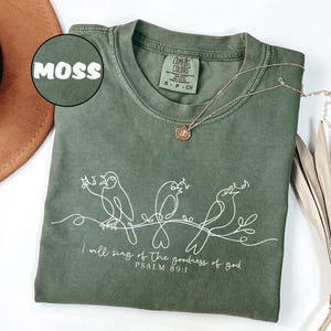 May include: A moss green t-shirt featuring a white line drawing of three birds on a branch, with the text "I will sing of the goodness of god, Psalm 89:1". A gold necklace with a pendant is placed on the shirt. The shirt is a size S-P-CH.