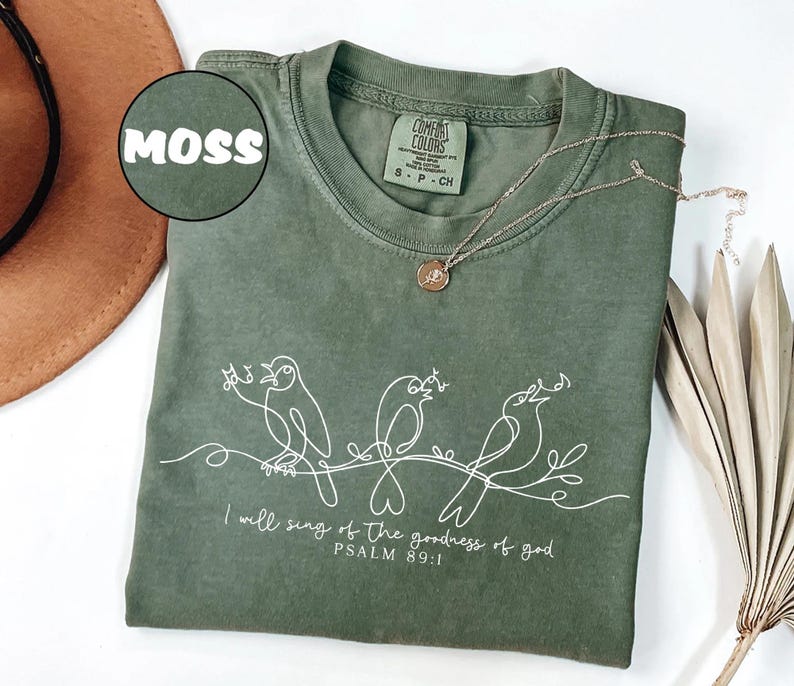 May include: A moss green t-shirt featuring a white line drawing of three birds on a branch, with the text "I will sing of the goodness of god, Psalm 89:1". A gold necklace with a pendant is placed on the shirt. The shirt is a size S-P-CH.