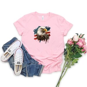 May include: A pink t-shirt with a graphic of a bald eagle with an American flag in the background. The eagle is facing the left side of the image. The flag is faded and has a paint splatter effect.