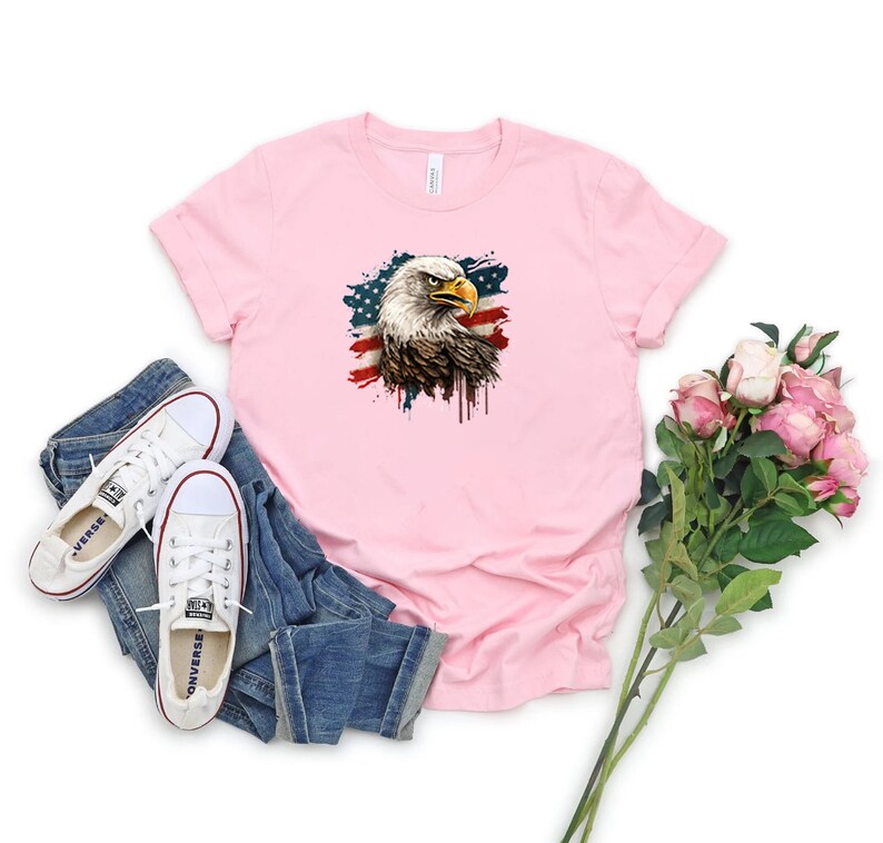 May include: A pink t-shirt with a graphic of a bald eagle with an American flag in the background. The eagle is facing the left side of the image. The flag is faded and has a paint splatter effect.