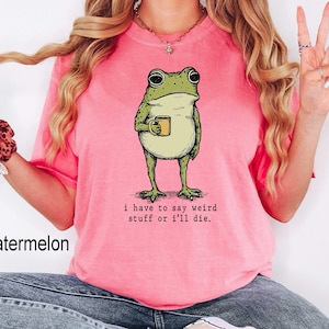 May include: A watermelon-colored t-shirt featuring a whimsical illustration of a frog holding a mug. The frog is green and beige, with the text "I have to say weird stuff or I'll die." printed below the frog.