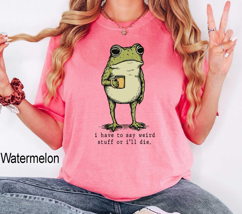 May include: A watermelon-colored t-shirt featuring a whimsical illustration of a frog holding a mug. The frog is green and beige, with the text "I have to say weird stuff or I'll die." printed below the frog.