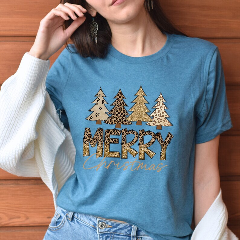 May include: A teal blue t-shirt with a graphic of four Christmas trees in a leopard print and gold glitter design. The text "Merry Christmas" is written in gold glitter letters.