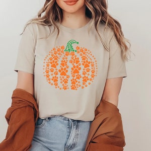 Peut inclure: T-shirt beige avec un motif de citrouille orange compos&eacute; d'empreintes de pattes et d'une tige verte. Le t-shirt est &agrave; col rond et &agrave; manches courtes. Le motif est sur le th&egrave;me de l'automne.