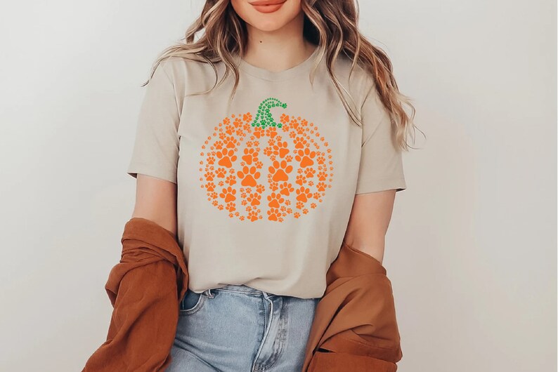 Peut inclure: T-shirt beige avec un motif de citrouille orange compos&eacute; d'empreintes de pattes et d'une tige verte. Le t-shirt est &agrave; col rond et &agrave; manches courtes. Le motif est sur le th&egrave;me de l'automne.