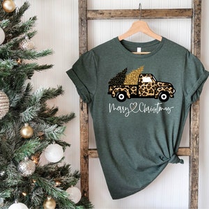 Op de afbeelding: Groen T-shirt met een afbeelding van een vrachtwagen die een kerstboom vervoert die is versierd met luipaardprint en gouden glitter. De tekst "Merry & Christmas" is onder de vrachtwagen gedrukt.