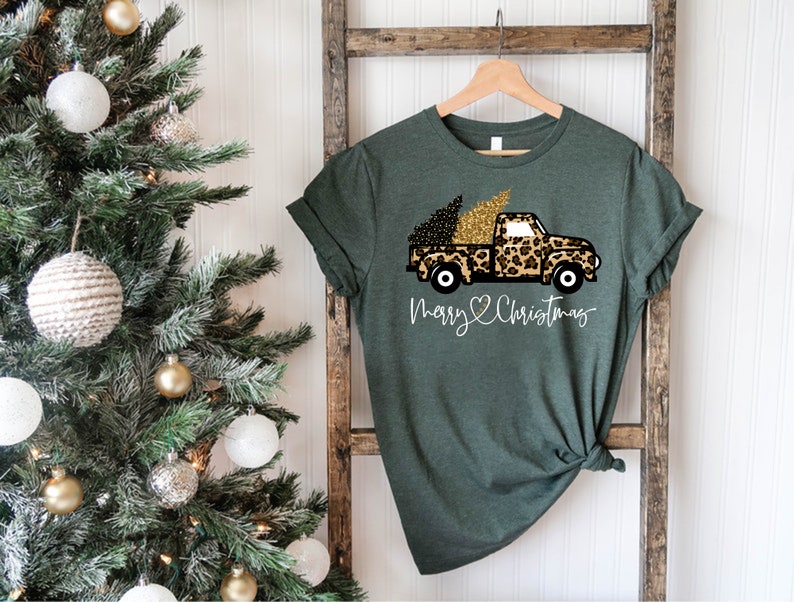 Op de afbeelding: Groen T-shirt met een afbeelding van een vrachtwagen die een kerstboom vervoert die is versierd met luipaardprint en gouden glitter. De tekst "Merry & Christmas" is onder de vrachtwagen gedrukt.