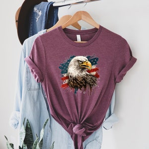 May include: A maroon t-shirt with a graphic of a bald eagle with an American flag background. The eagle's head is facing right and the flag is behind it. The flag is red, white, and blue with a white star pattern.