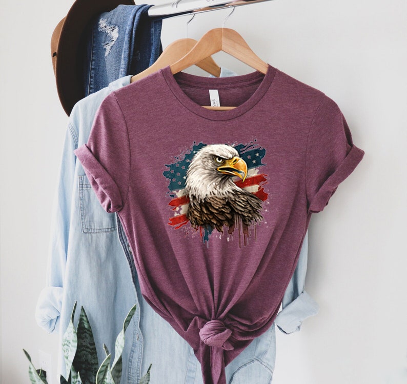 May include: A maroon t-shirt with a graphic of a bald eagle with an American flag background. The eagle's head is facing right and the flag is behind it. The flag is red, white, and blue with a white star pattern.
