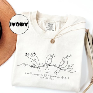 May include: Ivory Comfort Colors t-shirt with a black line drawing of three birds on a branch. The text "I will sing of the goodness of god, Psalm 89:1" is printed below. A gold necklace is draped across the shirt. The shirt is a small size.