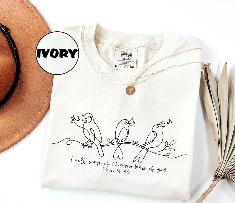 May include: Ivory Comfort Colors t-shirt with a black line drawing of three birds on a branch. The text "I will sing of the goodness of god, Psalm 89:1" is printed below. A gold necklace is draped across the shirt. The shirt is a small size.