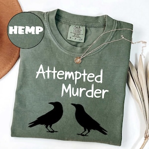 May include: Olive green t-shirt with the text "Attempted Murder" and two black raven silhouettes. The word "HEMP" is in a circle in the upper left corner. A gold necklace with a pendant is on the shirt.