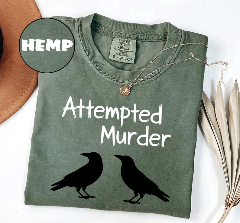 May include: Olive green t-shirt with the text "Attempted Murder" and two black raven silhouettes. The word "HEMP" is in a circle in the upper left corner. A gold necklace with a pendant is on the shirt.