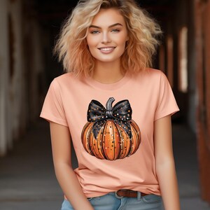 May include: A peach coloured t-shirt with a black and gold pumpkin with a black bow. The pumpkin has gold glitter and black stars on it.