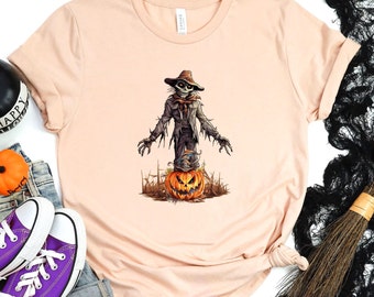 May include: A peach-colored t-shirt with a graphic of a spooky scarecrow standing on a jack-o'-lantern. The scarecrow is wearing a brown hat and a black jacket.