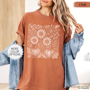 May include: A terracotta-colored t-shirt with a white floral and pumpkin design. The graphic features large daisies, leaves, and two pumpkins. The shirt has short sleeves and a crew neck. The text "MORE COLOR OPTIONS" is in a white bubble. The word "YAM" is in a brown rectangle.
