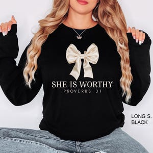 Puede incluir: Sudadera negra de manga larga con un lazo blanco y el texto "SHE IS WORTHY PROVERBS 31" en blanco.
