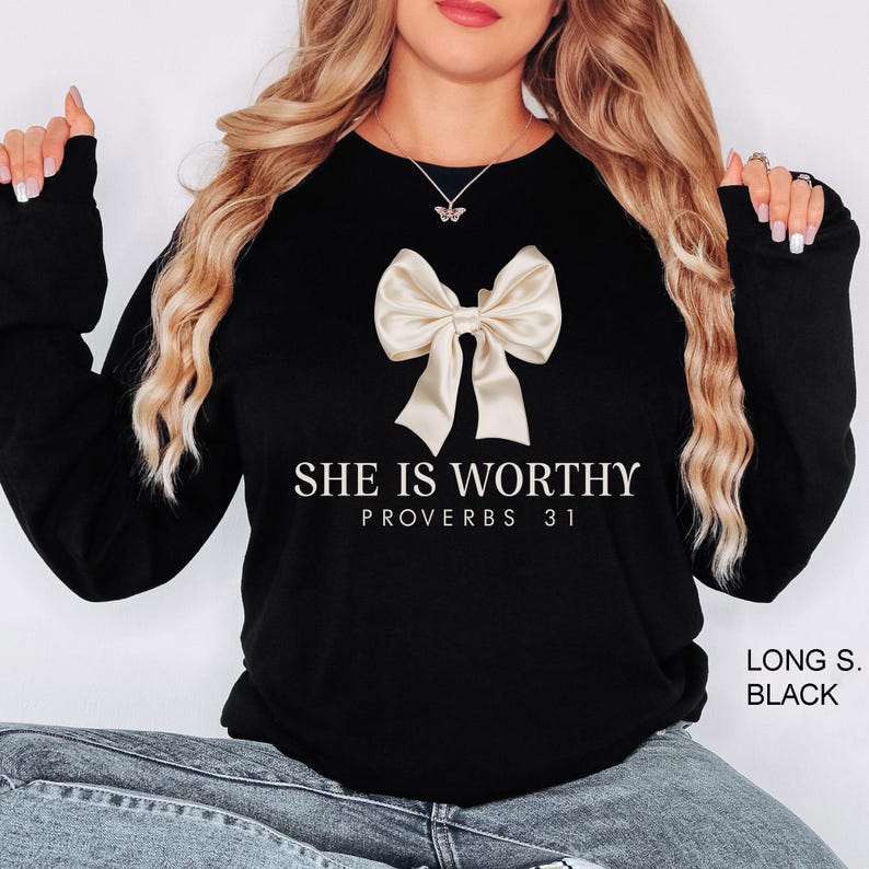 Puede incluir: Sudadera negra de manga larga con un lazo blanco y el texto "SHE IS WORTHY PROVERBS 31" en blanco.