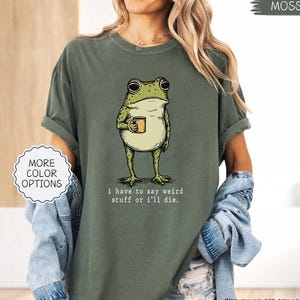 May include: Moss green t-shirt featuring a cartoon frog holding a mug. The frog is green and white with large eyes. The text "I have to say weird stuff or I'll die" is below the frog. The shirt has rolled-up sleeves.