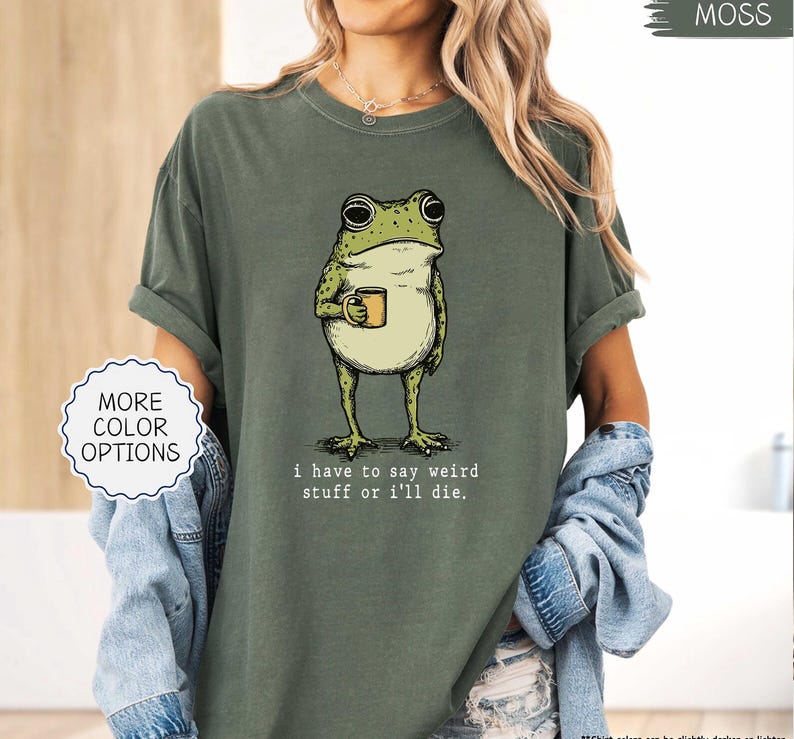 May include: Moss green t-shirt featuring a cartoon frog holding a mug. The frog is green and white with large eyes. The text "I have to say weird stuff or I'll die" is below the frog. The shirt has rolled-up sleeves.