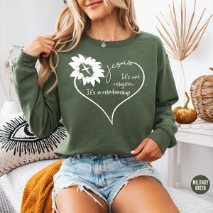 May include: A military green long-sleeve sweatshirt featuring a white graphic of a sunflower with a cross in the center, shaped into a heart outline. The text reads: Jesus It's not religion... It's a relationship. The sweatshirt has a crew neckline.