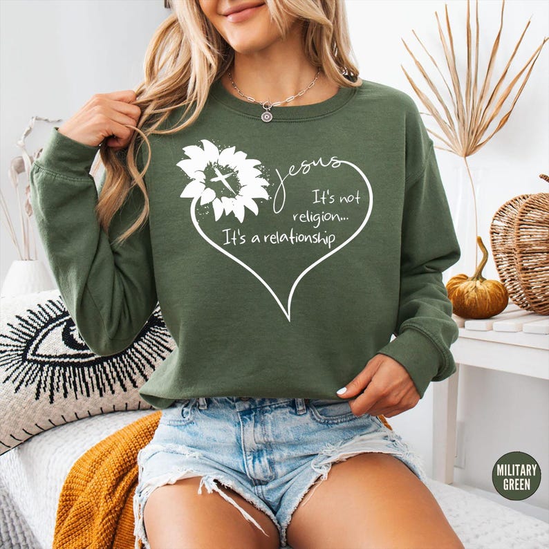 May include: A military green long-sleeve sweatshirt featuring a white graphic of a sunflower with a cross in the center, shaped into a heart outline. The text reads: Jesus It's not religion... It's a relationship. The sweatshirt has a crew neckline.
