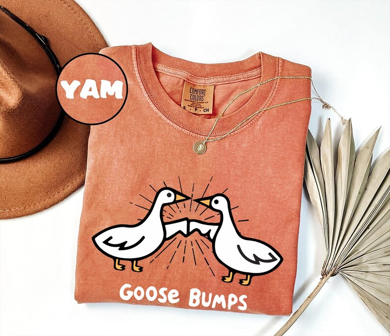 May include: A burnt orange t-shirt with a white graphic of two geese facing each other with the text "Goose Bumps" below. The shirt has a tag that says "Comfort Colors" and the word "YAM" is written in a circle on the top left corner of the image.