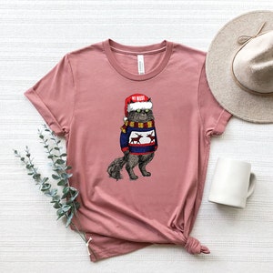 K&ouml;nnte beinhalten: Ein pinkfarbenes T-Shirt mit einem Aufdruck eines Katers, der einen Weihnachtsmann-Hut und einen Weihnachtspullover mit Rentieren tr&auml;gt. Der Text auf dem Hut lautet "My Woof".