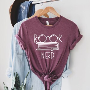 Pu&ograve; includere: Una maglietta color granata con testo bianco che recita "Book Nerd" e una grafica di un libro con gli occhiali sopra.